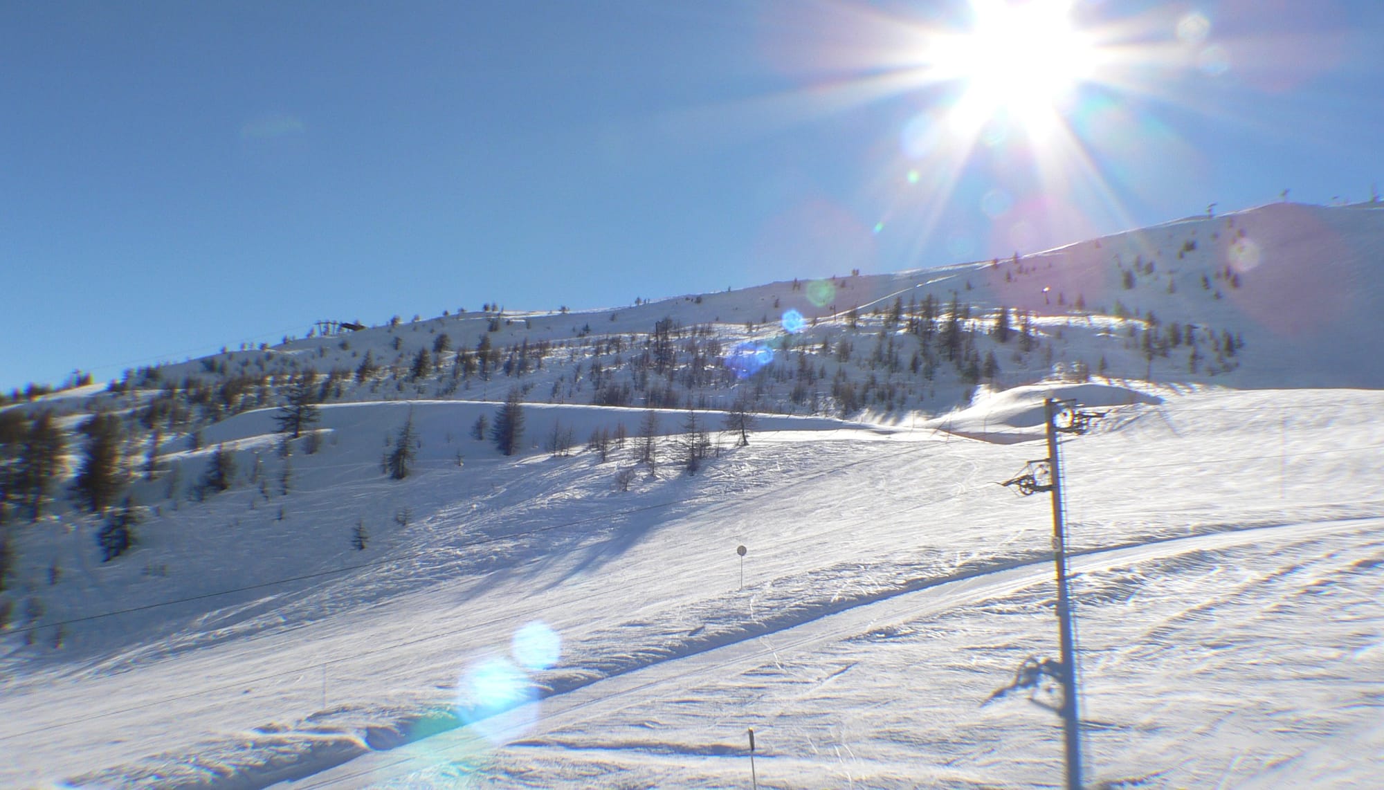 The image shows a sunny day at a ski resort, with a ski lift visible on the right side of the frame. The slopes are covered in snow, and there are tracks from skiers and snowboards.